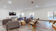This single-floor communal space is designed with a clean, contemporary aesthetic that promotes social interaction and relaxation. The open-concept layout is anchored by light wood-look flooring, crisp white walls, and recessed lighting throughout, creating a bright and welcoming atmosphere.

The main lounge area is generously proportioned, featuring multiple seating arrangements with plush gray armchairs, a sectional sofa accented with yellow and neutral throw pillows, and distinctive natural wood block side tables. A large wall-mounted flat-screen television is paired with a sleek, low-profile media console, making it an ideal space for group viewing. Potted indoor palms add a touch of greenery to the neutral color palette.

Adjacent to the lounge, a corridor-style seating nook displays bold yellow abstract artwork, providing a stylish transition between areas. This hallway leads to glass-panel doors that offer a sense of openness while defining the space.

A dedicated recreation room rounds out the space, equipped with a ping pong table and a shuffleboard table, alongside additional lounge seating. Windows throughout the space bring in natural light and exterior views. The overall design balances functionality with modern style, making it well-suited as a shared amenity space for residents or occupants.