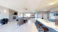 This single-floor space showcases an open-concept layout designed for comfortable socializing and entertaining. The vaulted ceiling rises prominently overhead, giving the interior a generous sense of height and airiness, while a ceiling fan with wood-toned blades adds both function and a subtle design detail. Light-toned, large-format tile flooring runs throughout, contributing to a clean and cohesive aesthetic.

A striking stone accent wall anchors the lounge area, housing a fireplace that serves as a focal point for the seating arrangement. Neutral-toned upholstered chairs and sofas are arranged thoughtfully around this area, creating an inviting conversational setting. A wall-mounted flat-screen television completes the lounge corner.

Adjacent to the lounge, a long quartz-topped bar counter with industrial-style bar stools provides an additional gathering spot, complemented by a sink and ample counter space. Beyond the seating area, a dining zone near the windows allows natural light to filter into the space, brightening the interior.

The walls feature a two-tone paint treatment in soft blue-gray and white, lending a polished, contemporary character throughout. The overall ambiance is welcoming, modern, and well-suited for communal use.