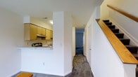 This two-story residence offers a practical and well-organized layout designed for comfortable everyday living. The main level features a semi-open kitchen, partially separated from the main living area by a structural dividing wall. The kitchen is equipped with light wood cabinetry, a range with an overhead exhaust fan, and a sink, providing ample workspace and storage. The countertop wraps around to create a functional pass-through area that connects the kitchen to the adjacent living space.

The main level flows into a carpeted hallway that leads to additional rooms, including a door-accessible space at the end of the corridor. The neutral carpet throughout adds warmth and a cohesive feel to the interior. Crisp white walls keep the space feeling clean and open, while recessed ceiling vents ensure proper climate control throughout.

A staircase with a warm wood handrail connects the main level to the upper floor, adding a classic residential touch to the design. The overall color palette is neutral and understated, with white walls, light wood accents, and soft grey carpeting working together to create a calm, move-in-ready environment. The layout prioritizes functionality, making efficient use of the available space across both floors.