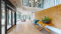 This contemporary lobby and common area is thoughtfully designed to make a striking first impression. The expansive entrance features large-format tile flooring and soaring double-height ceilings, creating an airy, open atmosphere throughout. A dramatic chandelier of globe pendant lights anchors the main lobby corridor, complemented by geometric starburst light fixtures that add a sculptural quality to the space.

Floor-to-ceiling glass curtain walls line the perimeter, flooding the interior with abundant natural light and framing panoramic views of the surrounding cityscape and skyline. The lounge area is furnished with an eclectic mix of colorful modular seating in teal, purple, yellow, and orange tones, arranged to encourage both relaxation and social interaction.

A standout textured wood accent wall adds warmth and visual depth, offset by lush tropical plants and a sleek round wooden table. A fully enclosed glass-walled meeting room provides a private yet transparent space for small gatherings, featuring a round table, modern chairs, and vibrant striped accent wallcovering.

Toward the rear, a refined elevator lobby finished in grey tile and warm vertical wall sconces houses dual stainless steel elevators. A small kitchenette or service alcove with a wall-mounted display screen rounds out the amenities, making this a well-appointed, design-forward communal space.