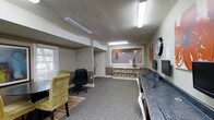 This single-floor space is designed to serve as a practical and well-organized business center or leasing office. The main area is finished with neutral-toned walls and carpeted flooring, creating a professional yet welcoming atmosphere. Fluorescent lighting is installed throughout, ensuring consistent, bright illumination across the entire space.

Along one wall, a continuous dark countertop workstation accommodates multiple computer terminals and peripherals, providing ample workspace for business tasks. Seating options, including a rolling office chair and an accent chair with a patterned fabric, add comfort and flexibility to the workspace.

A built-in shelving unit with organized cubby-style storage bins anchors the far wall, offering practical organization for documents and supplies. Adjacent to it, a pass-through service window with a white wainscoting surround connects to a secondary room, suggesting a reception or administrative function. A wall-mounted clock and decorative artwork add modest character to the otherwise utilitarian setting.

A window with horizontal blinds on the side wall allows for natural light to filter into the space. The textured ceiling and structural column contribute to the layout's defined zones, making the overall environment efficient and purposefully arranged for both staff and visitor use.