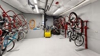 Located at 2715 California Ave SW in Seattle's West Seattle neighborhood, this dedicated indoor bicycle storage room offers a compact yet efficiently organized space for cyclists. The room features painted concrete walls and a smooth, light-colored concrete floor, giving it a clean and utilitarian appearance.

The space is outfitted with multiple red wall-mounted bicycle racks that accommodate bikes in a vertical orientation, maximizing the available floor area. Additional floor-level rail stands provide further storage flexibility for a variety of bicycle types and sizes. A security camera is mounted near the ceiling, providing surveillance coverage of the room's contents.

A utility corner enhances the room's functionality, featuring a wall-mounted air compressor with both coiled and full-length orange hoses, ideal for tire inflation and basic bike maintenance. Overhead, exposed ductwork, plumbing pipes, and fluorescent lighting fixtures line the ceiling, contributing to the room's practical, no-frills aesthetic.

Access is provided through a sturdy red metal door. The overall layout is thoughtfully arranged to allow residents to store and retrieve bicycles with ease, making this space a valuable amenity for cycling-oriented residents of the building.