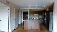 This single-floor space centers around an open-concept kitchen designed for both functionality and style. The kitchen is anchored by a substantial island with a granite countertop, providing ample workspace and serving as a natural divider between the kitchen and surrounding living areas. Dark espresso-toned cabinetry lines the walls, offering generous storage, while a decorative mosaic tile backsplash in warm earth tones adds texture and visual interest between the upper and lower cabinets. Stainless steel appliances, including a range and sink, complete the kitchen's modern, cohesive aesthetic. Warm hardwood flooring extends throughout the space, lending continuity and warmth. Track lighting mounted on the ceiling provides directed illumination across the kitchen area. A hallway visible from the kitchen leads to additional rooms, with multiple doors suggesting separate private spaces beyond. The overall layout feels open and practical, with clean lines and a neutral color palette that creates a welcoming, move-in-ready environment suited to everyday living.