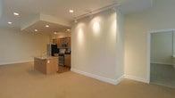 This single-floor space showcases a clean, modern design with an open-concept layout that promotes a sense of spaciousness and ease of movement. The living area features neutral-toned carpeting and crisp white walls, creating a bright and welcoming atmosphere. A sleek track lighting system runs across the ceiling, providing adjustable, directional illumination throughout the main living space.

The kitchen is thoughtfully integrated into the open floor plan, separated by a peninsula-style counter with a built-in sink — ideal for casual dining or meal preparation. Warm wood-toned cabinetry lines the kitchen walls, complemented by stainless steel appliances including a refrigerator, gas range, and microwave. Recessed ceiling lights in the kitchen add a layer of functional brightness to the cooking area.

The overall color palette — soft beiges, warm woods, and clean whites — lends the space a calm, understated elegance. The seamless transition between the carpeted living zone and the kitchen area enhances the open feel of the layout. The minimalist aesthetic and practical design make this a functional and inviting space suited for everyday living.