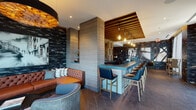 This single-floor space is thoughtfully designed as a upscale lounge and co-working environment, blending social and functional areas within a cohesive, contemporary aesthetic. The centerpiece is a long, teal-tiled bar counter lined with navy blue and gold-accented bar chairs, offering ample seating for both work and leisure. A striking slatted wood ceiling element floats above the bar area, fitted with Edison-style pendant lights that cast a warm, ambient glow throughout the space.

The floors throughout are finished in a rich herringbone-patterned tile, lending a sophisticated texture underfoot. Dark, marbled stone-effect wall cladding anchors the design, complemented by large-scale black-and-white artwork that adds visual drama. A comfortable lounge area features a tufted cognac leather sofa alongside plush teal seating, creating an inviting social zone.

Toward one end of the space, floor-to-ceiling glass doors open to an outdoor terrace, flooding the interior with natural light and providing seamless indoor-outdoor connectivity. A flat-screen display and workstations integrated into the bar counter make the space equally suited for productive use. The overall ambiance is refined and moody, balancing warmth and modernity throughout.
