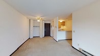 This single-floor unit offers a straightforward, functional layout designed to maximize usable space. The main living area is finished with neutral wall-to-wall carpeting and white walls, creating a clean, versatile backdrop. A ceiling fan with a light fixture is centrally positioned overhead, providing both air circulation and ambient lighting throughout the space.

Adjacent to the living area, a built-in closet with bi-fold doors offers convenient storage near the entryway. The dark-toned front door serves as a clear focal point at the entry, contributing a contrast against the otherwise light interior palette. Dark baseboard trim runs throughout, adding subtle definition to the floors and walls.

The kitchen is set apart by a half-wall partition, creating a semi-open pass-through design that maintains a visual connection to the living area. The kitchen features warm-toned wood cabinetry, light countertops, and under-cabinet appliances. A dedicated overhead light fixture illuminates the kitchen workspace efficiently. The overall ambiance of the unit is practical and understated, suited to those seeking a low-maintenance, move-in-ready space.