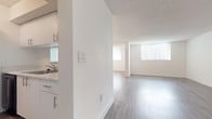 This single-floor unit is designed with a clean, minimalist aesthetic throughout, featuring a predominantly white color palette and light-toned wood-style flooring across the main living areas. The open-concept layout connects a spacious living zone to a defined dining area, the latter highlighted by a pendant light fixture hanging overhead. A small pass-through window on a partial wall creates a visual link between the kitchen and the living space, adding architectural interest while maintaining a sense of openness.

The galley-style kitchen is efficiently laid out, featuring white cabinetry with sleek bar-pull hardware, a light stone-finish countertop, a single-basin sink, and black appliances including a refrigerator, range, and dishwasher. An adjacent closet with a bi-fold door provides convenient storage, and a washer unit is also located in this area.

The bedroom is finished with plush carpeting and benefits from a sliding glass door fitted with vertical blinds, allowing natural light to fill the room while offering access to an outdoor space with green views beyond.

Throughout the unit, textured ceilings, recessed HVAC vents, and white trim detailing contribute to a cohesive and well-maintained interior. The overall ambiance is bright, airy, and move-in ready.