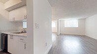 This single-floor unit is designed with a clean, minimalist aesthetic throughout, featuring a predominantly white color palette and light-toned wood-style flooring across the main living areas. The open-concept layout connects a spacious living zone to a defined dining area, the latter highlighted by a pendant light fixture hanging overhead. A small pass-through window on a partial wall creates a visual link between the kitchen and the living space, adding architectural interest while maintaining a sense of openness.

The galley-style kitchen is efficiently laid out, featuring white cabinetry with sleek bar-pull hardware, a light stone-finish countertop, a single-basin sink, and black appliances including a refrigerator, range, and dishwasher. An adjacent closet with a bi-fold door provides convenient storage, and a washer unit is also located in this area.

The bedroom is finished with plush carpeting and benefits from a sliding glass door fitted with vertical blinds, allowing natural light to fill the room while offering access to an outdoor space with green views beyond.

Throughout the unit, textured ceilings, recessed HVAC vents, and white trim detailing contribute to a cohesive and well-maintained interior. The overall ambiance is bright, airy, and move-in ready.