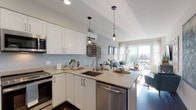 This thoughtfully designed single-floor residence showcases a contemporary aesthetic with a warm, coastal-inspired color palette throughout. The open-concept layout seamlessly connects the kitchen and living area, creating an inviting and functional space ideal for both everyday living and entertaining.

The kitchen is outfitted with crisp white shaker-style cabinetry, taupe quartz countertops, and a full suite of stainless steel appliances, including a range, over-the-range microwave, dishwasher, and refrigerator. A peninsula with bar seating and pendant lighting adds both functionality and style, while dark hardwood flooring grounds the space with warmth and contrast.

The living area flows naturally from the kitchen and is bathed in natural light from large sliding glass doors that open to an outdoor balcony, offering pleasant views of the surrounding neighborhood. The space features soft gray walls and recessed lighting, contributing to a calm and airy atmosphere.

The two bedrooms are generously proportioned, each featuring large windows that bring in abundant natural light and scenic outdoor views. Neutral gray walls, dark hardwood flooring, and ample space define each room. The full bathroom is well-appointed with a soaking tub and shower combination featuring marble-style tile surround, white cabinetry, and a broad vanity mirror.