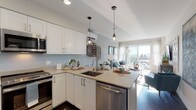 This thoughtfully designed single-floor residence showcases a contemporary aesthetic with a warm, coastal-inspired color palette throughout. The open-concept layout seamlessly connects the kitchen and living area, creating an inviting and functional space ideal for both everyday living and entertaining.

The kitchen is outfitted with crisp white shaker-style cabinetry, taupe quartz countertops, and a full suite of stainless steel appliances, including a range, over-the-range microwave, dishwasher, and refrigerator. A peninsula with bar seating and pendant lighting adds both functionality and style, while dark hardwood flooring grounds the space with warmth and contrast.

The living area flows naturally from the kitchen and is bathed in natural light from large sliding glass doors that open to an outdoor balcony, offering pleasant views of the surrounding neighborhood. The space features soft gray walls and recessed lighting, contributing to a calm and airy atmosphere.

The two bedrooms are generously proportioned, each featuring large windows that bring in abundant natural light and scenic outdoor views. Neutral gray walls, dark hardwood flooring, and ample space define each room. The full bathroom is well-appointed with a soaking tub and shower combination featuring marble-style tile surround, white cabinetry, and a broad vanity mirror.