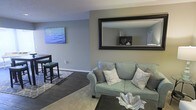 This single-floor residence offers a thoughtfully designed living space that combines modern style with everyday comfort. The living area serves as the heart of the home, featuring cool gray walls that create a calm, neutral backdrop for the carefully curated furnishings. A plush sage green sofa anchors the seating area, accented with decorative throw pillows in coordinating patterns and tones. A large, bold-framed wall mirror adds depth and visual interest, reflecting the room and amplifying the sense of space. Dark hardwood-style flooring grounds the room, complemented by a patterned area rug that defines the living zone. A small bistro-style table with matching stools creates an intimate dining or workspace nook, adding versatility to the layout. A vibrant ocean-themed canvas artwork brings a pop of color to the walls, contributing to the relaxed, contemporary ambiance. The open layout encourages a natural flow between areas, making the space feel connected and spacious. Overall, the residence presents a clean, modern aesthetic with warm, welcoming details throughout.
