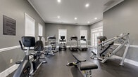 Located at 135 Reynoldsburg New Albany Road South in Reynoldsburg, Ohio, this fitness center is a thoughtfully designed single-floor facility offering a full range of exercise equipment in a refined, functional environment. The space is finished with neutral gray walls accented by crisp white crown molding and chair rail trim, lending a clean and polished aesthetic throughout.

The fitness center is divided into distinct workout zones. The cardio area features multiple Landice treadmills and a True elliptical trainer, positioned to take advantage of natural light streaming through white-trimmed windows and glass-paneled French doors. The strength training section is equally well-appointed, housing a Hoist cable machine, adjustable weight benches, a Cybex recumbent bike, and a full dumbbell rack with a variety of weights. A large wall-mounted mirror expands the visual depth of the space while supporting proper form during training.

Recessed lighting provides consistent, even illumination throughout, complemented by a wall-mounted flat-screen television for entertainment during workouts. Dark rubber flooring provides durability and safety underfoot. A wall clock and posted fitness center guidelines complete the practical appointments of the space, contributing to an organized and welcoming environment suited for a wide range of fitness routines.