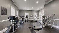 Located at 135 Reynoldsburg New Albany Road South in Reynoldsburg, Ohio, this fitness center is a thoughtfully designed single-floor facility offering a full range of exercise equipment in a refined, functional environment. The space is finished with neutral gray walls accented by crisp white crown molding and chair rail trim, lending a clean and polished aesthetic throughout.

The fitness center is divided into distinct workout zones. The cardio area features multiple Landice treadmills and a True elliptical trainer, positioned to take advantage of natural light streaming through white-trimmed windows and glass-paneled French doors. The strength training section is equally well-appointed, housing a Hoist cable machine, adjustable weight benches, a Cybex recumbent bike, and a full dumbbell rack with a variety of weights. A large wall-mounted mirror expands the visual depth of the space while supporting proper form during training.

Recessed lighting provides consistent, even illumination throughout, complemented by a wall-mounted flat-screen television for entertainment during workouts. Dark rubber flooring provides durability and safety underfoot. A wall clock and posted fitness center guidelines complete the practical appointments of the space, contributing to an organized and welcoming environment suited for a wide range of fitness routines.