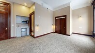 Located at 888 Delaware Ave in Buffalo, NY, this one-bedroom, one-bathroom unit blends historic architectural character with modern updates. The spacious bedroom is the heart of the unit, featuring plush neutral carpeting, rich dark wood trim and baseboards, elegant crown molding, and a decorative fireplace with a classic mantel — a charming nod to the building's heritage. A large double-hung window with dark wood casing floods the room with natural light and offers views of the surrounding neighborhood. Wall sconces with brass accents provide warm ambient lighting throughout. The unit entry features a set of stately dark wood double doors with a transom detail, adding a refined, classic touch. A compact kitchenette with gray cabinetry, a mini refrigerator, and a microwave offers convenient in-unit functionality. The bathroom is thoughtfully appointed with large-format marble-look porcelain tile floors, a full walk-in shower with floor-to-ceiling marble tile and sliding glass doors, a vanity with chrome fixtures, and a large mirror. Dark wood door frames throughout tie the design together cohesively, creating a space that balances timeless architectural detail with contemporary finishes.