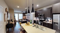 This single-floor space showcases a thoughtfully designed open-concept layout where the kitchen and living area connect seamlessly, creating an inviting and functional environment. The kitchen is anchored by a large island with a light-toned countertop, offering seating for casual dining and additional prep space. Three striking matte black pendant lights hang above the island, serving as a bold focal point. Dark espresso cabinetry lines the kitchen walls, complemented by a dark tile backsplash and a full suite of stainless steel appliances, including an electric range, microwave, and under-counter storage. A deep single-basin sink with a modern faucet adds to the kitchen's practical appeal. Rich dark hardwood flooring flows throughout, tying the kitchen and living areas together cohesively. The living space is comfortably furnished and benefits from natural light streaming through large windows framed with blinds and drapery. A ceiling fan overhead enhances both comfort and airflow. Recessed lighting throughout provides consistent illumination, while the overall aesthetic balances contemporary design with a warm, livable ambiance.