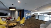 Located at 201 S Elliot Rd in Chapel Hill, NC, this single-floor commercial office space combines modern design with a professional, welcoming atmosphere. The interior is defined by a bold color palette, featuring striking deep-blue accent walls that contrast with neutral gray tones and warm herringbone-patterned carpeting throughout. A well-appointed lounge area greets visitors with contemporary seating, including sculptural white chairs and vibrant yellow swivel chairs set around low, organic-shaped tables — creating an inviting space for casual meetings or collaboration. Overhead, a large drum pendant light anchors the lounge area, complemented by recessed ceiling lighting and a distinctive circular soffit detail that adds architectural interest. Curated wall art adorns the blue accent walls, lending the space a polished, creative character. Adjacent to the lounge, an open-plan work area features individual workstations with partitioned cubicles, providing a functional and focused environment for day-to-day operations. A glass-paneled partition helps define zones within the open floor plan while maintaining a sense of transparency and connectivity. The overall ambiance is professional yet approachable, making this space well-suited for a dynamic office environment.