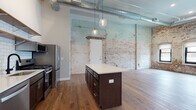 Located at 150 N 3rd St in the heart of downtown Columbus, Ohio, this urban loft seamlessly blends historic industrial character with modern finishes. The open-concept living area commands attention with soaring ceilings, exposed ductwork, and original whitewashed brick walls that lend warmth and texture throughout the space. Rich hardwood flooring flows continuously across the main living areas, grounding the loft's expansive feel.

The kitchen is a standout feature, anchored by a large center island with a quartz countertop and dark cabinetry. A full suite of stainless steel appliances — including a gas range, dishwasher, and French door refrigerator — is complemented by classic white subway tile backsplash and open wood shelving. Pendant globe lights illuminate the workspace with an industrial elegance. In-unit laundry is conveniently located adjacent to the kitchen.

The bedroom area benefits from the same exposed brick detailing and hardwood flooring, with arched windows offering natural light and views of the surrounding downtown streetscape. A walk-in closet with wire shelving organization provides generous storage. The bathroom features warm hardwood flooring, a walk-in shower with clean white tile surround, and a vanity with ample counter space, delivering a crisp, functional finish.