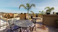 Located at 840 17th St in San Diego's vibrant East Village neighborhood, this rooftop terrace offers an impressive open-air retreat with sweeping panoramic views of the downtown San Diego skyline and surrounding cityscape. The space is thoughtfully designed to maximize outdoor living, featuring a combination of composite wood decking and artificial turf that creates distinct zones for relaxation and entertainment.

A dedicated lounge area is outfitted with cushioned chaise lounges, perfect for soaking up the abundant Southern California sunshine. Nearby, an ornate cast-iron bistro table and chair set provides an inviting outdoor dining nook framed by tall, swaying palm trees that lend a tropical ambiance. A built-in grill station makes the terrace well-suited for al fresco entertaining.

Decorative iron railings line the perimeter, ensuring safety while preserving unobstructed sightlines across the urban horizon. Potted plants and mature palms are thoughtfully placed throughout, adding lush greenery against the bright blue sky. A charming stucco access structure with a red-tiled roof and arched doorway serves as the entry point to the terrace, adding a touch of Mediterranean architectural character. Classic-style lamp posts complete the refined, welcoming aesthetic of this exceptional rooftop outdoor space.