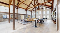Located at Solace Pond View in Colorado Springs, CO, this single-floor community clubhouse offers a generously sized gathering space defined by dramatic architectural character. The centerpiece of the building is the expansive living room, where soaring vaulted ceilings are supported by an impressive exposed timber truss system with rich dark wood beams and vertical posts. Natural light pours in through a series of large windows positioned at multiple heights along the walls, creating a bright and welcoming atmosphere throughout.

The flooring features a distinctive combination of large-format stone tile and wood-look planks arranged in a patterned layout, adding visual depth to the open floor plan. A standout design element is the bold gabion stone accent wall — wire-framed panels filled with natural rock — that anchors the dining and lounge areas with an earthy, textural contrast to the white walls.

The living area is furnished with comfortable seating arrangements, a wall-mounted television, and recreational amenities including a billiards table and foosball table. A dedicated office space features a glass-walled conference room with a wood table, upholstered seating, and a mounted display screen. A closet rounds out the layout. Views through the windows reveal the surrounding outdoor amenities and neighborhood, further connecting the interior to its Colorado setting.