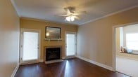 This single-story space centers around a welcoming living area that combines classic design elements with comfortable functionality. The room features rich, dark hardwood floors that extend throughout, reflecting light and adding warmth to the space. A tile-surround fireplace serves as a natural focal point, complemented by a simple white mantel shelf and a wall-mounted mirror above, enhancing the sense of depth in the room. Crown molding frames the ceiling, adding a refined architectural detail, while a textured ceiling with a central ceiling fan provides both style and practical air circulation. Natural light flows generously through multiple windows fitted with white blinds, flanking the fireplace on either side and brightening the interior throughout the day. The warm yellow-toned walls contribute to the inviting atmosphere, while crisp white baseboards provide a clean contrast against the dark flooring. The overall ambiance is cozy yet polished, offering a versatile living space that balances classic architectural touches with a relaxed, livable feel.