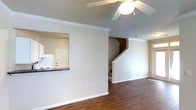 This two-story residence offers a thoughtfully designed layout spread across two levels connected by a staircase visible from the main living area. The main floor features an open living space with warm hardwood-style flooring, crown molding, and a ceiling fan that adds both functionality and a finishing touch to the room. Vaulted or angled ceiling lines lend a sense of added volume to the space. A pass-through window connects the kitchen to the living area, promoting an open feel while maintaining a degree of separation between the two spaces. The kitchen, visible through the pass-through, includes dark countertops and standard appliances, including a refrigerator and range. Neutral-toned walls throughout create a clean, versatile backdrop suitable for a variety of interior styles. White trim and baseboards provide crisp contrast against the warm flooring. The staircase, framed with white balusters and a wood handrail, leads to the second floor, where additional living spaces are located. Overall, the home presents a functional and straightforward layout with classic design details.