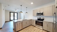This single-floor unit presents a clean, contemporary design throughout. The open-concept kitchen serves as the centerpiece, equipped with light wood-tone cabinetry, granite countertops, a subway tile backsplash, and a full suite of stainless steel appliances including a range, microwave, refrigerator, and dishwasher. An L-shaped peninsula with an undermount sink and pendant lighting overhead creates a natural division between the kitchen and living area, while light wood-look plank flooring ties the two spaces together seamlessly.

The living area is bright and open, featuring a ceiling fan and a set of dark-framed French doors that provide access to an outdoor space and allow natural light to fill the room. The kitchen also benefits from views through these doors, enhancing the overall sense of openness.

The bedrooms feature neutral carpet flooring and windows with blinds that frame outdoor views. A generously sized walk-in closet with multi-tier wire shelving provides substantial storage capacity. The bathroom includes a granite-topped vanity with light wood cabinetry, a tiled tub and shower combination, and matching plank flooring, maintaining a consistent, modern aesthetic throughout the unit.