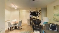 This single-floor space showcases an open-concept design that seamlessly connects the living, dining, and kitchen areas into one cohesive environment. The light hardwood flooring runs throughout, lending warmth and visual continuity across the space.

The kitchen is a standout feature, boasting dark cabinetry that contrasts sharply against a light tile backsplash and stainless steel appliances, including a refrigerator. A peninsula with bar seating provides both a functional prep area and a casual dining option, illuminated by sleek pendant lighting and track lighting above.

Adjacent to the kitchen, the dining area comfortably accommodates a round table with upholstered chairs, creating an intimate setting for meals. The living area is anchored by a bold leather accent chair and a patterned area rug, adding texture and personality to the space.

Ceiling fans and layered lighting — including track fixtures and table lamps — ensure the space is well-lit and comfortable. The neutral wall tones and thoughtful décor, including a decorative wall-mounted sculpture, contribute to the space's modern yet inviting ambiance.