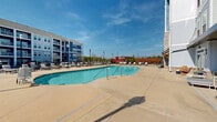 Located at 3010 Furr Court in Charlotte, NC, this generously sized outdoor amenity space offers a well-appointed resort-style experience for residents. At its centerpiece sits a large, freeform swimming pool with crystal-clear blue water, accessible via stainless steel entry rails and flanked by an expansive concrete sundeck. The sundeck is lined with ample lounge chairs, providing plenty of space for sunbathing and relaxation beneath open Carolina skies.

Surrounding the pool area, the space is thoughtfully divided into distinct social zones. A dedicated fire pit seating area features Adirondack-style chairs arranged around a raised brick fire feature, perfect for cooler evenings. String lights overhead create a warm, inviting atmosphere throughout the patio. A wood pergola anchors an al fresco dining zone outfitted with multiple table-and-chair groupings, ideal for outdoor gatherings.

Iron fencing encloses the entire space, offering both security and an open sightline to the broader community. Classic lamp post lighting is distributed throughout, ensuring the area remains functional and attractive after dark. The multi-story residential building, finished with a combination of brick, siding, and large windows, frames the backdrop of the outdoor space, with ground-level glazing connecting interior common areas directly to the pool deck. A cornhole game adds a playful recreational touch to the overall amenity-rich environment.
