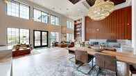 This expansive single-floor space is a masterclass in contemporary interior design, offering a generous and open layout that accommodates a variety of social and functional zones. The grand entrance lobby immediately impresses with soaring double-height ceilings and a dramatic wall of floor-to-ceiling black-framed windows, flooding the interior with abundant natural light and offering views of the surrounding exterior. Comfortable lounge seating, layered rugs, and artful pendant lighting create a welcoming atmosphere from the moment you enter.

Moving deeper into the space, the main gathering area showcases a striking combination of materials and textures — light-toned wide-plank hardwood floors transition seamlessly into ornate encaustic-style patterned tile, grounding the dining area with a bold visual anchor. A solid wood dining table surrounded by leather cantilever chairs sits beneath sculptural woven chandelier pendants. A sleek marble-topped bar counter, set against a backdrop of dark stone tile cladding, adds a refined and functional focal point.

Accent walls clad in rich terracotta-toned tiles and dramatic natural stone panels contribute warmth and depth throughout. The hallway, adorned with cascading stacked pendant lights in organic forms, connects the various zones with artistic continuity, lined with artwork and framed by the same cohesive hardwood flooring. Wooden slatted ceiling details and structural columns complete the refined, curated aesthetic of this remarkable space.