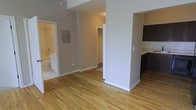 This single-floor space offers a thoughtfully arranged open layout that connects the main living area to the kitchen, bathroom, and bedroom corridor with ease. Warm-toned hardwood flooring extends throughout the space, lending a cohesive and inviting feel to each area.

The main living area is open and airy, framed by neutral gray-toned walls and clean white trim and baseboards. A structural column subtly defines the transition between the living space and the kitchen zone without fully enclosing either area.

The kitchen features dark cabinetry with a contrasting light stone or tile backsplash, creating a modern aesthetic. Dark lower cabinets are paired with upper storage, providing functional organization space.

A hallway leads to a walk-in closet, offering generous storage with visible shelving. The bathroom, accessible from the main living area, is finished in light tones with white cabinetry, a vanity, and a bathtub, giving the space a clean, classic look.

An electrical panel is neatly recessed into the wall along the hallway. Overall, the space maintains a consistent, minimal design language with neutral colors, hardwood floors, and practical layouts that make each area functional and welcoming.