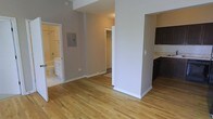 This single-floor space offers a thoughtfully arranged open layout that connects the main living area to the kitchen, bathroom, and bedroom corridor with ease. Warm-toned hardwood flooring extends throughout the space, lending a cohesive and inviting feel to each area.

The main living area is open and airy, framed by neutral gray-toned walls and clean white trim and baseboards. A structural column subtly defines the transition between the living space and the kitchen zone without fully enclosing either area.

The kitchen features dark cabinetry with a contrasting light stone or tile backsplash, creating a modern aesthetic. Dark lower cabinets are paired with upper storage, providing functional organization space.

A hallway leads to a walk-in closet, offering generous storage with visible shelving. The bathroom, accessible from the main living area, is finished in light tones with white cabinetry, a vanity, and a bathtub, giving the space a clean, classic look.

An electrical panel is neatly recessed into the wall along the hallway. Overall, the space maintains a consistent, minimal design language with neutral colors, hardwood floors, and practical layouts that make each area functional and welcoming.