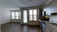This single-floor space features an open-concept layout that seamlessly connects the kitchen and living areas. The dark hardwood flooring runs throughout, providing a warm, grounding contrast to the light-colored walls and trim. Natural light is a defining characteristic of this space, with multiple large windows fitted with horizontal blinds lining the walls of the living area. A glass-paned door leads to what appears to be a balcony or outdoor area, further enhancing the sense of openness and connection to the exterior.

The kitchen is positioned along one wall and is equipped with stainless steel appliances, including a range and an over-the-range microwave. Dark upper cabinetry pairs with granite or stone-look countertops, offering a modern aesthetic. Recessed and track lighting in the ceiling provides additional illumination to complement the abundant natural light. The open flow between the kitchen and the adjacent living area creates a functional and cohesive layout, making the space feel spacious and well-connected. Overall, the design emphasizes clean lines, modern finishes, and a bright, airy atmosphere.