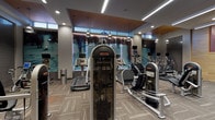 This contemporary fitness center is thoughtfully designed to deliver a premium workout experience across a spacious, open floor plan. The cardio zone is anchored by a row of Star Trac treadmills, ellipticals, and stair climbers positioned beneath a striking large-scale photographic mural and high clerestory windows that draw in natural light. Warm wood-paneled ceiling insets contrast with sleek grey tones and recessed linear lighting, lending the space a polished, upscale ambiance. Pendant-style industrial fixtures add a distinctive design accent overhead. The strength training area features a power rack, a comprehensive free weight section with organized plate storage, and cable machines, all set against large mirrors that visually expand the space. Patterned carpet tile with orange accent lines defines the main floor, while a dedicated functional training and yoga area offers wood flooring, medicine balls, and wall-mounted accessories. A built-in wood-framed shelving unit near the entrance displays informational materials and fitness resources, complemented by smart control panels. The facility also includes a decorative tile accent wall and glass-railed staircase, contributing to its refined, modern aesthetic throughout.