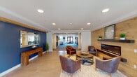 This well-appointed single-floor community space offers a cohesive blend of comfort and contemporary design across several distinct areas. The central lounge serves as the heart of the space, featuring a striking wood-paneled accent wall with a sleek linear fireplace, surrounded by plush leather armchairs and a round coffee table atop a patterned area rug — creating a warm and inviting gathering spot. Recessed lighting illuminates the crisp white ceilings throughout, while light hardwood-style flooring ties the common areas together seamlessly.

Adjacent to the lounge, a sunlit sitting room commands attention with expansive floor-to-ceiling windows that frame outdoor views of a furnished patio and surrounding landscape. This area is furnished with a generous sectional sofa, accent chairs, a wall-mounted television, and a light wood console — ideal for casual relaxation.

A large, open multipurpose room with neutral carpeting and built-in reception counters offers flexible space for events or gatherings, with direct sightlines to the lounge beyond. Throughout the space, curated artwork in navy and gold tones adds a refined coastal aesthetic. Lush potted greenery and thoughtful accent pieces complete the polished, welcoming atmosphere of this community facility.