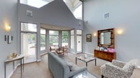 This single-story leasing center makes a strong first impression with its dramatic vaulted ceiling, which draws the eye upward and creates an expansive, airy atmosphere throughout the space. Large skylights and oversized windows flood the interior with natural light, enhancing the open and inviting feel of the room. The main seating area is furnished with a plush gray sofa and a glass-top coffee table, offering a comfortable and professional setting for prospective residents. A curved bay window enclosure at the far end of the space features a small table and chairs, providing an intimate spot bathed in natural light with views of the surrounding landscaped grounds and pool area. A wooden credenza along one wall displays informational materials and is accented by a decorative framed mirror and wall sconce, adding warmth and character to the otherwise neutral gray-toned interior. Neutral carpeting and white trim work complement the light gray walls, creating a clean, cohesive aesthetic. Direct exterior access is available through a glass-paneled door, seamlessly connecting the interior to the outdoor amenity areas.