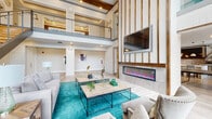 This contemporary clubhouse is designed to impress, anchored by a soaring double-height main lobby that immediately commands attention. Floor-to-ceiling windows flood the space with natural light while offering views of the outdoor pool area, creating a seamless connection between the interior and exterior. A sleek steel-and-wood staircase leads to an open upper mezzanine level, framed by a glass-panel railing that adds an airy, modern touch.

The main lounge area is elegantly appointed with neutral upholstered seating arranged around wood-topped coffee tables atop a vibrant teal area rug. A striking floor-to-ceiling accent wall features vertical wood slat detailing, a wall-mounted flat-screen TV, and a long linear electric fireplace with colorful flame display, creating a bold focal point. Exposed wood ceiling beams overhead add warmth and architectural character throughout.

The fully equipped catering kitchen offers generous quartz countertops, warm wood cabinetry with a diamond-pattern wine rack accent, a tiled backsplash, stainless steel appliances, and a large granite-topped island — ideal for entertaining. Adjacent to the lounge, a dedicated games room provides a pool table, ping-pong table, and wall-mounted TV, with direct access to the outdoor area. Light-toned large-format tile flooring unifies the space throughout.