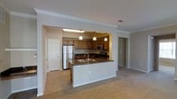 This single-floor residence offers a thoughtfully designed open-concept layout that seamlessly connects the kitchen and living areas, creating a spacious and functional environment. The kitchen is a standout feature, showcasing warm wood cabinetry, dark granite countertops, and a full suite of stainless steel appliances, including a refrigerator, microwave, and range. A peninsula island with a double sink and an overhanging granite countertop provides additional prep space and serves as a natural divider between the kitchen and the open living area. Pendant lighting hangs above the island, adding a refined touch to the space. Crown molding runs along the ceiling throughout, lending an elegant, finished look to the interior. The living area features neutral-toned carpeting that complements the warm beige walls, creating a cohesive and inviting ambiance. A separate entry door is visible from the main living space, contributing to a practical and well-organized floor plan. The consistent use of neutral tones, quality finishes, and architectural detailing throughout makes this a polished and comfortable single-floor living space.
