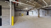 This single-level enclosed parking garage offers a well-organized, functional space designed to accommodate a variety of vehicles, including full-size trucks. The structure is built with concrete masonry block walls and a poured concrete floor, giving the space a solid, durable character. Overhead, an exposed concrete ceiling carries a network of piping, conduit, and mechanical systems, including a fire suppression system fitted with red-accented components. Fluorescent and surface-mounted lighting fixtures provide consistent illumination throughout the interior.

The garage is divided into clearly marked parking stalls, with painted numbering on the floor to designate individual spaces. Dedicated accessible parking stalls are also present, identified with standard ADA markings. Yellow bollards are strategically positioned throughout to protect structural columns and utility areas from vehicle impact.

Access to the garage is provided through a sectional glass-panel overhead garage door, alongside a secondary interior entry door finished in black. Small windows along the perimeter walls allow limited natural light to supplement the artificial lighting. A designated bicycle storage area with wall-mounted hooks is located within the space. The overall environment is utilitarian and well-maintained, prioritizing practicality and organized vehicle and equipment storage.