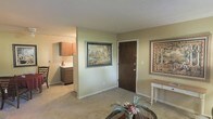 This single-floor residence offers a comfortable and well-organized layout designed for everyday living. The living area serves as the heart of the space, featuring neutral-toned carpeting that adds warmth underfoot and soft, light green walls that create a calm and inviting atmosphere. Decorative framed artwork, including nature-themed tapestry-style pieces, adorn the walls and contribute a classic, homey character to the interior. A glass-topped accent table with a floral arrangement provides an elegant focal point near the entryway. The solid dark wood front door anchors the entry area, lending a sense of privacy and security. A console table with turned legs is positioned along one wall, offering a functional yet decorative touch. Adjacent to the living area, the kitchen is partially visible and features dark wood cabinetry with ample storage, a countertop workspace, and practical overhead cabinet units. The kitchen's warm wood tones complement the overall color palette of the home. A dining chair can be glimpsed near the kitchen, suggesting a dedicated dining area nearby. Throughout the space, the design emphasizes understated comfort, with cohesive finishes and traditional décor elements that give the home a lived-in, welcoming feel.