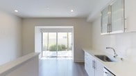 This single-story space showcases a clean, contemporary design characterized by a bright and airy aesthetic. The interior is finished with light-toned hardwood-style flooring that runs throughout, complementing the neutral warm-white walls and crisp white trim. Recessed lighting in the smooth white ceiling provides even illumination across the open layout.

The kitchen area is outfitted with modern white cabinetry featuring brushed metal hardware, offering generous counter space with a light-colored solid surface countertop and a stainless steel sink. Upper cabinetry with frosted or glass-panel doors adds a refined touch to the overall design. A kitchen island or peninsula provides additional workspace and visual definition within the open floor plan.

A large sliding glass door serves as a prominent architectural feature, flooding the interior with an abundance of natural light and offering a direct view of a private outdoor courtyard. The outdoor space, framed by white walls and mature vegetation including palm trees and shrubs, enhances the sense of a seamless indoor-outdoor connection. The overall ambiance is calm, minimal, and well-lit, making the space feel welcoming and versatile.