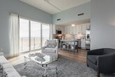 Located steps from the Virginia Beach shoreline on Atlantic Avenue, this single-floor coastal retreat is thoughtfully designed with a light, airy aesthetic throughout. Soft blue-gray walls, warm light wood flooring, and an abundance of natural light from large windows create a cohesive and calming atmosphere.

The kitchen is well-appointed with white shaker cabinetry, a herringbone tile backsplash, stainless steel appliances, and a freestanding blue kitchen island with a white countertop — a vibrant coastal accent that doubles as a casual dining space with acrylic bar seating.

The living area features elevated ceilings, large windows with ocean-facing views, and an open layout that fosters a bright, relaxed ambiance. Coastal décor including blue-toned rugs and sheer white curtains enhance the beachside character.

The bedroom is generously sized with dual windows capturing views of the surrounding Virginia Beach skyline and neighboring structures, allowing for exceptional natural light. A walk-in closet with built-in rod storage and shelving provides ample organization space.

The bathroom is finished with classic black-and-white mosaic tile flooring, a glass-enclosed walk-in shower with white subway tile surround, and a white vanity, delivering a clean, timeless coastal aesthetic throughout.
