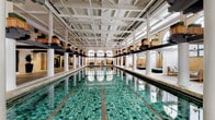 This impressive single-level indoor aquatic facility showcases a compelling blend of historic architectural character and refined amenity design. At its centerpiece sits a generously sized lap pool lined with vivid green mosaic tiles, flanked on both sides by wide poolside promenades featuring intricate, multicolored geometric tilework in earthy tones of amber, slate, and sage. Soaring ceilings supported by bold steel beams and white cylindrical columns lend a grand, industrial-meets-classical atmosphere throughout the space.

Arched windows trimmed with decorative blue tile allow natural light to filter in along the exposed brick walls, reinforcing the building's historic character. Along the pool's perimeter, sheer-curtained cabana areas house cushioned lounge daybeds, providing semi-private relaxation spaces directly poolside. Elevated planters with greenery accent the structural beams overhead, adding a natural touch to the interior.

Tucked away from the main pool hall are intimate lounge alcoves finished in pale brick with arched inlays of deep teal arabesque tile. These seating areas are furnished with teak-framed sofas and rustic wooden coffee tables, creating cozy retreats. The entry area is marked by tall potted trees flanking glass-paneled doors, setting an inviting tone upon arrival.