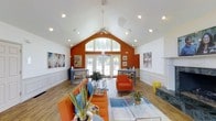 This lively and welcoming single-floor space makes an immediate impression with its dramatic vaulted ceilings that soar to a high peak, creating an expansive, airy atmosphere. The bold orange accent wall serves as a striking focal point, lending warmth and energy to the interior while contrasting beautifully against the crisp white walls and ceiling. A ceiling fan anchors the apex of the vaulted ceiling, enhancing both form and function. Natural light pours generously into the space through a large arched window and a set of French doors at the far end, offering views of lush greenery outside and seamlessly connecting the interior to the outdoors. The main living area is thoughtfully arranged with vibrant orange seating, a sleek glass coffee table, and colorful artwork that adds personality and visual interest throughout. Wainscoting detail along the lower walls adds a classic architectural touch that balances the space's contemporary energy. Recessed lighting and track lights are strategically placed along the ceiling to complement the natural light. A pair of workstation-style tables flank the French doors, making the space as functional as it is inviting. Warm wood-toned flooring ties the entire design together, creating a cohesive and dynamic environment.