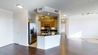This 2-bedroom, 2-bathroom residence offers a generous, thoughtfully designed single-floor layout. The open-concept living and kitchen area serves as the heart of the home, anchored by rich, dark hardwood floors that extend throughout the main living spaces. The kitchen is well-equipped with stainless steel appliances, including a French door refrigerator with ice and water dispenser, a built-in microwave, and a range. Warm honey-toned cabinetry provides ample storage, complemented by dark granite countertops that add a sleek contrast. A peninsula with an integrated sink and breakfast bar seating connects the kitchen to the living area, enhancing the space's functionality and flow. Track lighting illuminates the kitchen, while flush-mount ceiling fixtures brighten the surrounding areas. A wide hallway visible from the main living space leads to the bedrooms and additional rooms, suggesting a practical and well-organized floor plan. The overall ambiance balances modern finishes with warm tones, creating a comfortable and inviting atmosphere throughout.