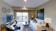 This single-floor living space showcases a thoughtfully designed interior that balances contemporary aesthetics with functional comfort. The main living area is anchored by a plush white sofa adorned with decorative throw pillows and a cozy faux fur blanket, complemented by a charcoal accent chair that adds depth to the seating arrangement. A bold, large-scale portrait painting in dark tones serves as a striking focal point on the wall, lending an artistic and sophisticated atmosphere to the room.

The space benefits from abundant natural light, courtesy of a full-width sliding glass door that opens onto a private balcony. The outdoor area features a bistro-style table and chairs, framed by potted topiaries, offering a pleasant outdoor retreat with a lush green view beyond the railing. Neutral-toned walls and recessed ceiling lighting create a warm, welcoming ambiance throughout.

A black-and-white striped bowl coffee table sits atop a layered area rug, serving as a stylish centerpiece. A sleek media console along the opposite wall provides additional functionality. The overall design reflects a modern, curated sensibility with carefully chosen décor elements.