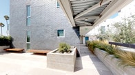 Located at 3008 Santa Monica Blvd in Santa Monica, California, this single-level outdoor space offers a thoughtfully designed courtyard environment that blends modern architecture with carefully curated natural elements. The space is defined by a striking contrast of materials — charcoal gray brick exterior walls, smooth concrete planters, and large-format light stone tile paving — creating a clean, contemporary aesthetic throughout.

Lush, drought-tolerant landscaping is a hallmark of the space, with ornamental grasses, bamboo, native shrubs, and river rock ground cover arranged in raised concrete planter beds. These plantings add texture, movement, and a sense of calm to the environment. Iconic Southern California palm trees are visible against the open sky, reinforcing the locale's character.

A covered walkway with an exposed metal trellis ceiling runs along the building's edge, providing a shaded passage between areas while maintaining an open, airy feel. Warm wood benches integrated into the concrete planters offer functional seating throughout. The open-air courtyard section benefits from abundant natural sunlight and open sky views, making it an inviting space for gathering or quiet relaxation. The overall ambiance is serene, modern, and distinctly California in character.