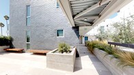 Located at 3008 Santa Monica Blvd in Santa Monica, California, this single-level outdoor space offers a thoughtfully designed courtyard environment that blends modern architecture with carefully curated natural elements. The space is defined by a striking contrast of materials — charcoal gray brick exterior walls, smooth concrete planters, and large-format light stone tile paving — creating a clean, contemporary aesthetic throughout.

Lush, drought-tolerant landscaping is a hallmark of the space, with ornamental grasses, bamboo, native shrubs, and river rock ground cover arranged in raised concrete planter beds. These plantings add texture, movement, and a sense of calm to the environment. Iconic Southern California palm trees are visible against the open sky, reinforcing the locale's character.

A covered walkway with an exposed metal trellis ceiling runs along the building's edge, providing a shaded passage between areas while maintaining an open, airy feel. Warm wood benches integrated into the concrete planters offer functional seating throughout. The open-air courtyard section benefits from abundant natural sunlight and open sky views, making it an inviting space for gathering or quiet relaxation. The overall ambiance is serene, modern, and distinctly California in character.