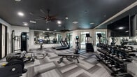Located at 1820 Brittmoore Rd in Houston, TX, this well-appointed fitness center delivers a comprehensive workout environment within a thoughtfully designed single-floor layout. The space makes a strong first impression with its striking dark ceiling, recessed lighting, and dual ceiling fans that keep air circulating throughout the room — a practical and stylish combination.

The flooring features a geometric, herringbone-patterned carpet tile in layered shades of gray, adding visual depth while providing a comfortable surface underfoot. Large windows along the perimeter flood the space with natural light and offer views of the surrounding greenery, creating an open and energizing atmosphere.

A full-length mirror wall visually expands the room and allows users to monitor their form during workouts. The cardio zone is equipped with treadmills, elliptical trainers, a stair climber, and Peloton spin bikes with integrated screens. The strength training area includes a cable machine, adjustable weight benches, battle ropes, and a well-stocked dumbbell rack ranging across multiple weight levels.

Entry is through sleek, black-framed glass sliding doors, reinforcing the modern aesthetic. The space is cleanly organized, with clear zones for different workout types, making it easy to navigate and use efficiently. Overall, this fitness center combines contemporary design with a practical, fully equipped exercise environment.