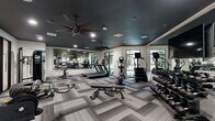 Located at 1820 Brittmoore Rd in Houston, TX, this well-appointed fitness center delivers a comprehensive workout environment within a thoughtfully designed single-floor layout. The space makes a strong first impression with its striking dark ceiling, recessed lighting, and dual ceiling fans that keep air circulating throughout the room — a practical and stylish combination.

The flooring features a geometric, herringbone-patterned carpet tile in layered shades of gray, adding visual depth while providing a comfortable surface underfoot. Large windows along the perimeter flood the space with natural light and offer views of the surrounding greenery, creating an open and energizing atmosphere.

A full-length mirror wall visually expands the room and allows users to monitor their form during workouts. The cardio zone is equipped with treadmills, elliptical trainers, a stair climber, and Peloton spin bikes with integrated screens. The strength training area includes a cable machine, adjustable weight benches, battle ropes, and a well-stocked dumbbell rack ranging across multiple weight levels.

Entry is through sleek, black-framed glass sliding doors, reinforcing the modern aesthetic. The space is cleanly organized, with clear zones for different workout types, making it easy to navigate and use efficiently. Overall, this fitness center combines contemporary design with a practical, fully equipped exercise environment.