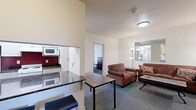 Located at 327 Eddy St in Ithaca, NY, this six-bedroom, two-bathroom unit is thoughtfully laid out across a single floor. The combined kitchen and living area serves as the heart of the unit, featuring dark granite-style countertops, white cabinetry, a coil-top electric range, and a built-in microwave set against a bold red accent wall. The living room offers carpeted flooring, two plush sofas, a wall-mounted flat-screen TV, and a central coffee table, creating a comfortable shared gathering space. A hallway connects the common areas to the private bedrooms and bathrooms. Each bedroom is equipped with carpeted flooring, a dresser, a desk with a chair, and individual climate control units. Several bedrooms benefit from large windows that invite natural light and provide views of surrounding greenery. The two full bathrooms are finished with light tile flooring, vanities with ample cabinet storage, large mirrors, and shower/tub combos with sliding glass doors — one bathroom features a double-sink vanity. The unit's straightforward layout and well-distributed room count make it well-suited for shared occupancy in the Ithaca area.